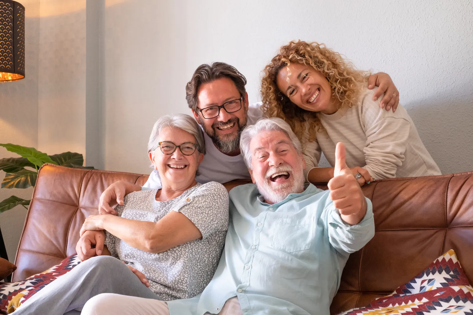 Four smiling family members embracing on a brown leather couch, older man giving thumbs up.